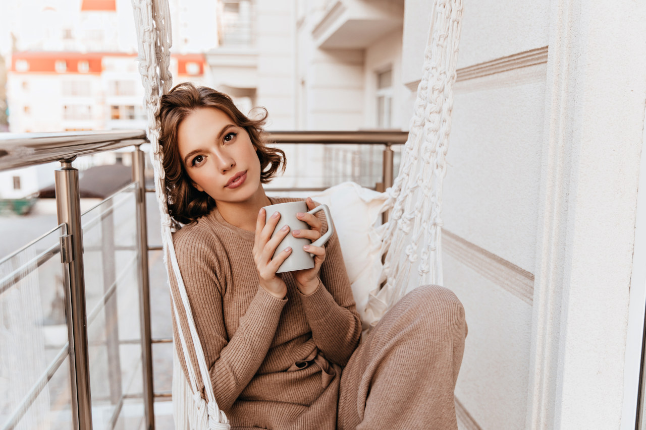 Beautiful girl in knitted dress drinking coffee in morning. Romantic caucasian young woman holding cup of tea at balcony.